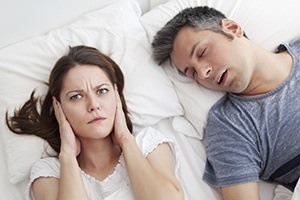 Woman covering ears next to snoring man in need of snoreguard sleep apnea therapy